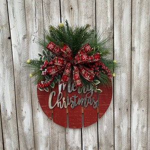 May include: A red wooden round sign with the words "Merry Christmas" in white lettering. The sign is decorated with a red and white plaid bow and green pine branches with red berries.