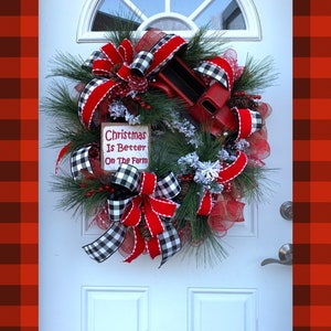 May include: A Christmas wreath with red and black buffalo plaid ribbon, red velvet ribbon, and a red toy truck. The wreath is decorated with pine branches, red berries, and faux snow. A sign on the wreath reads "Christmas Is Better On The Farm".
