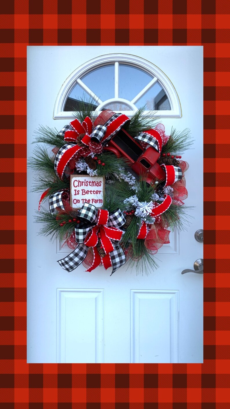 May include: A Christmas wreath with red and black buffalo plaid ribbon, red velvet ribbon, and a red toy truck. The wreath is decorated with pine branches, red berries, and faux snow. A sign on the wreath reads "Christmas Is Better On The Farm".