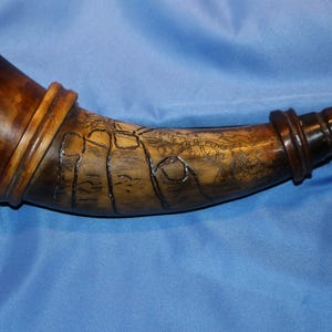 May include: A powder horn with a dark brown finish, featuring a map design etched onto its surface. The horn has a rounded base and a tapered spout, with decorative bands. The overall design suggests a historical or antique item.