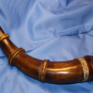 May include: A curved, dark brown horn with decorative gold-colored bands and accents. The horn has a bulbous end with a carved design and a small wooden knob. The horn is set against a blue background.