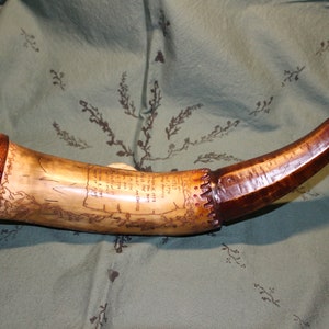 May include: A wooden and bone powder horn with a carved inscription and a wooden cap. The horn is decorated with a brown stain and has a metal tip.
