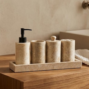 Op de afbeelding: Een badkamerset van beige steen, bestaande uit een zeepdispenser, twee cilindrische containers en een tandenborstelhouder, alles op een rechthoekige tray. De set staat op een houten wastafel, met een witte handdoek.