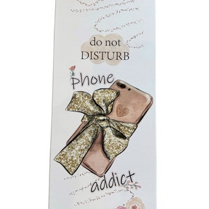 K&ouml;nnte beinhalten: Rechteckiges wei&szlig;es Schild mit einer Illustration eines ros&eacute;goldenen Telefons, einer goldglitzernden Schleife und den Worten "do not DISTURB" und "phone addict". Das Schild hat ein dekoratives Blumen- und Punktmuster.