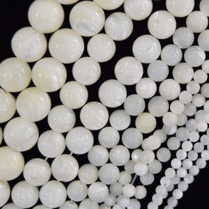 May include: A collection of round, white mother-of-pearl beads arranged in a gradient from large to small sizes. The beads have a smooth, iridescent surface with a natural luster, strung together on thin, clear filaments against a black background.
