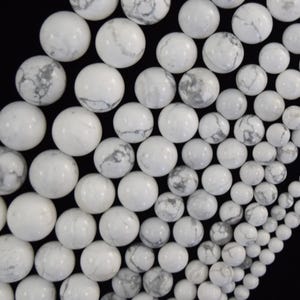 May include: A collection of round white howlite beads featuring natural grey veining patterns. The beads vary in size and are arranged in a dense cluster against a solid black background, showcasing a smooth, polished surface texture throughout.