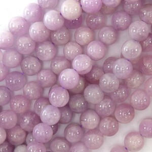 May include: A strand of pale lavender round gemstone beads. The beads are smooth and polished, with a translucent appearance.