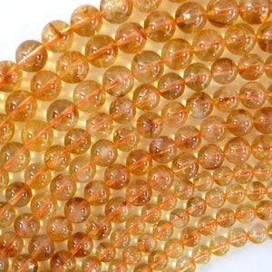 May include: A strand of round, translucent, golden yellow citrine beads with a slight orange hue. The beads have a smooth, polished finish and are strung on a clear elastic cord.