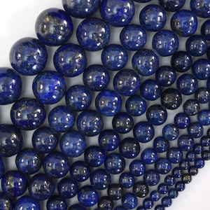 May include: A collection of round lapis lazuli beads in varying sizes, featuring a deep blue color with natural gold-colored pyrite inclusions. The smooth, polished spheres are arranged in a gradient pattern from large to small, creating a textured surface.