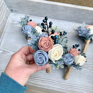 May include: A set of five boutonnieres with blue, white, and pink felt flowers, greenery, and blue berries. The boutonnieres are tied with twine.