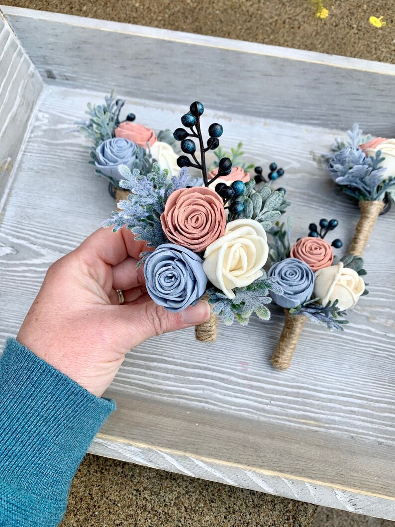 May include: A set of five boutonnieres with blue, white, and pink felt flowers, greenery, and blue berries. The boutonnieres are tied with twine.