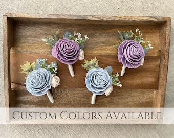 May include: Three wooden flower boutonnieres in a wooden tray. One boutonniere is lavender, one is light blue, and one is white. The tray has the text "Teton Wood Blooms" on it. CUSTOM COLORS AVAILABLE is written on the bottom of the tray.