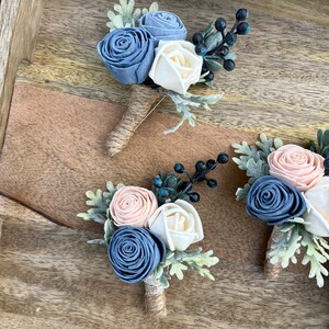 May include: Three boutonnieres with blue, white, and peach fabric flowers, greenery, and black berries. The boutonnieres are arranged on a wooden surface.