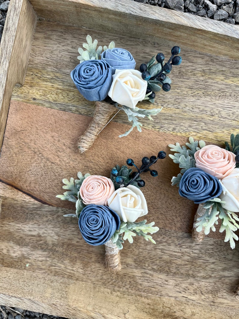 May include: Three boutonnieres with blue, white, and peach fabric flowers, greenery, and black berries. The boutonnieres are arranged on a wooden surface.