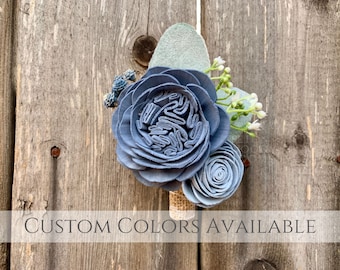 May include: A blue and white sola wood flower boutonniere with greenery. CUSTOM COLORS AVAILABLE.