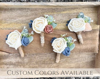 May include: Four wooden flower boutonnieres with white, peach, and blue flowers, greenery, and twine.  The boutonnieres are arranged on a wooden surface.  Custom colors available.