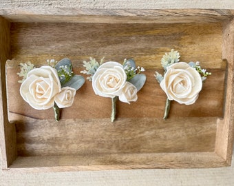 May include: Three ivory-colored sola wood flower boutonnieres with greenery accents. Each boutonniere features a larger rose and a smaller rose.