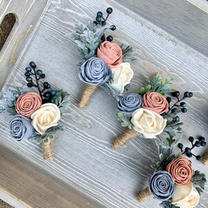 May include: Four boutonnieres with a rustic theme. Each boutonniere features three felt flowers in shades of dusty blue, blush pink, and ivory, with sprigs of greenery and blue berries.