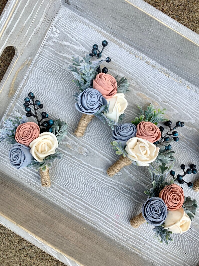 May include: Four boutonnieres with a rustic theme. Each boutonniere features three felt flowers in shades of dusty blue, blush pink, and ivory, with sprigs of greenery and blue berries.