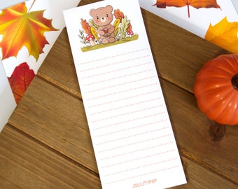 May include: A white notepad with a cute bear illustration holding a heart, surrounded by autumn leaves and mushrooms. The notepad has dotted lines for writing and the text "JOLLITYPOP" at the bottom. The notepad is on a wooden surface.