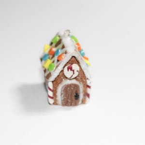 May include: A miniature gingerbread house ornament with a brown body, white icing, and colorful candy decorations. The roof is adorned with colorful candies, and the door has a heart-shaped detail. Red and white striped candy canes frame the house.