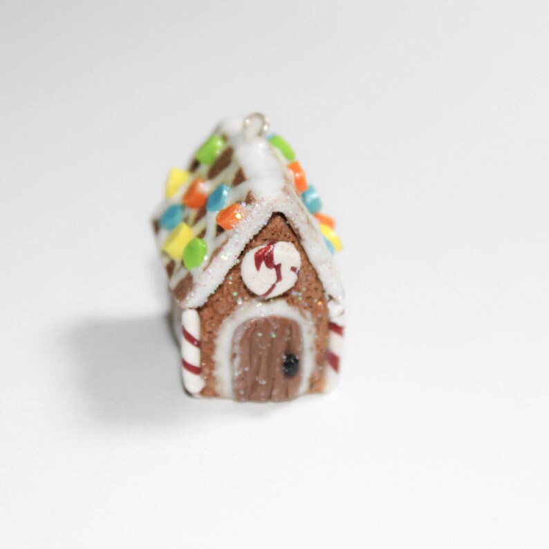May include: A miniature gingerbread house ornament with a brown body, white icing, and colorful candy decorations. The roof is adorned with colorful candies, and the door has a heart-shaped detail. Red and white striped candy canes frame the house.