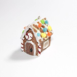 May include: A miniature gingerbread house ornament with a brown base, white icing, and colorful candy decorations. The roof is decorated with colorful candies, and the door has a red and white striped candy cane design. A small silver loop is attached for hanging.