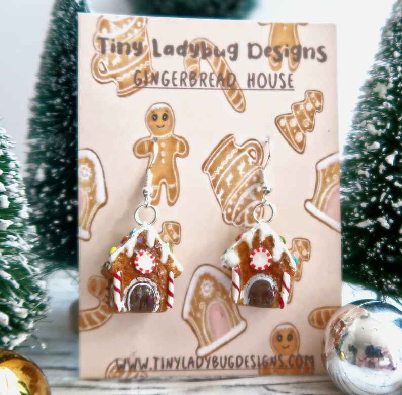May include: A pair of gingerbread house earrings with silver hooks. The earrings are brown with white icing, candy cane accents, and colorful sprinkles. The earrings are displayed on a card with gingerbread-themed illustrations and the text "Tiny Ladybug Designs GINGERBREAD HOUSE".