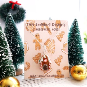 May include: A silver necklace with a gingerbread house pendant. The pendant is brown with white icing, red and white candy cane accents, and a dark brown door. The background features a pink card with gingerbread designs and the text "Tiny Ladybug Designs GINGERBREAD HOUSE".