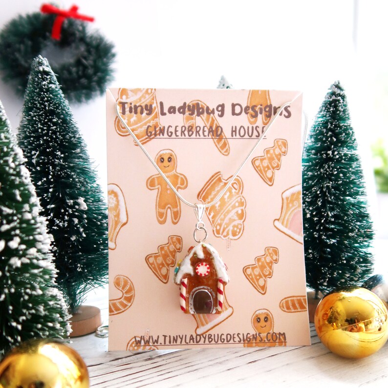 May include: A silver necklace with a gingerbread house pendant. The pendant is brown with white icing, red and white candy cane accents, and a dark brown door. The background features a pink card with gingerbread designs and the text "Tiny Ladybug Designs GINGERBREAD HOUSE".