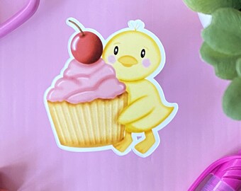 May include: A yellow chick sticker embracing a pink cupcake topped with a red cherry. The cupcake has a golden-brown base and pink icing. The chick has a yellow body, a yellow beak, and pink cheeks. The sticker is on a pink background.