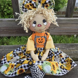 May include: A fabric doll with curly blonde yarn hair and a large bow featuring a black and white buffalo check pattern with sunflowers. The doll wears a yellow shirt under a dress with a sunflower, truck, and pumpkin print, plus black and white check legs.