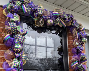 May include: A purple, gold, and green Mardi Gras wreath with gold and purple beads hanging from it. The wreath is decorated with bows and is hanging on a door.