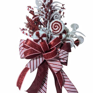May include: A red and white striped ribbon bow with glitter accents, topped with a white and red candy cane swirl, white and red candy canes, and white and red curly embellishments.