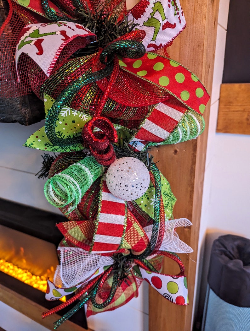May include: A Christmas wreath with red, green, and white ribbon, mesh, and a white ornament. The wreath is decorated with a green and white striped ribbon, a red and white striped ribbon, and a green and white polka dot ribbon.