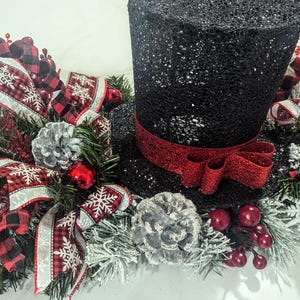 May include: A black glitter top hat with a red ribbon, surrounded by a festive arrangement. The arrangement includes red and black plaid ribbons, silver pinecones, red berries, and faux evergreen branches, creating a holiday centerpiece.