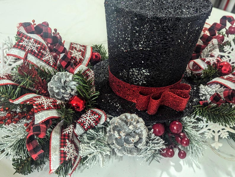 May include: A black glitter top hat with a red ribbon, surrounded by a festive arrangement. The arrangement includes red and black plaid ribbons, silver pinecones, red berries, and faux evergreen branches, creating a holiday centerpiece.