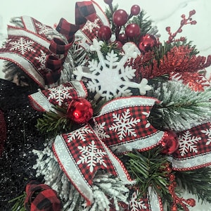 May include: A festive holiday wreath featuring a black glitter base, adorned with red and black buffalo check ribbons, white snowflake accents, and red ornaments. Greenery and red berries add to the winter theme.