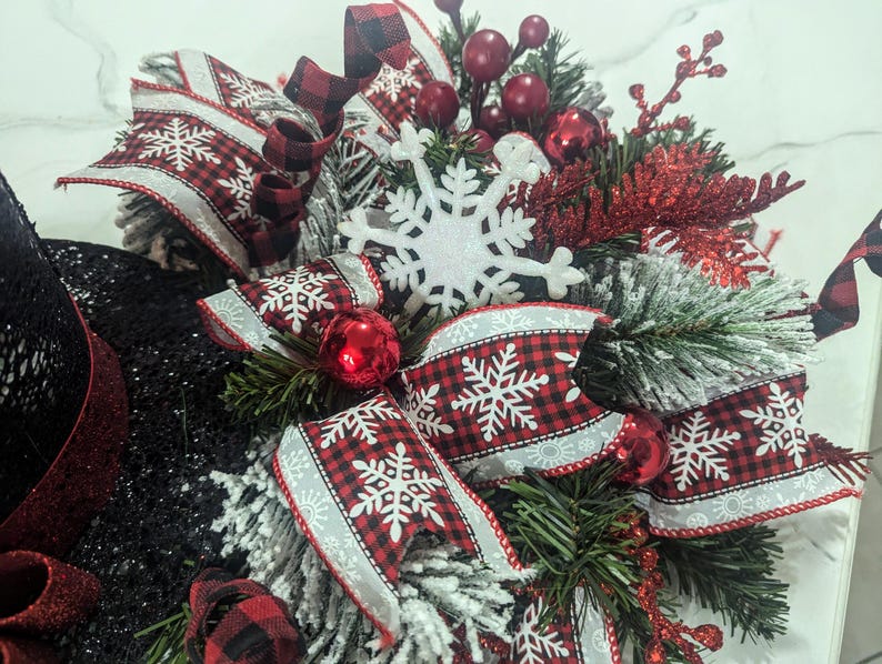 May include: A festive holiday wreath featuring a black glitter base, adorned with red and black buffalo check ribbons, white snowflake accents, and red ornaments. Greenery and red berries add to the winter theme.