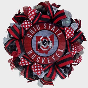 May include: A round wreath featuring the Ohio State Buckeyes logo in the center. The wreath is decorated with red, black, and white ribbons, including polka dot and striped patterns. The logo is on a wooden-style background.