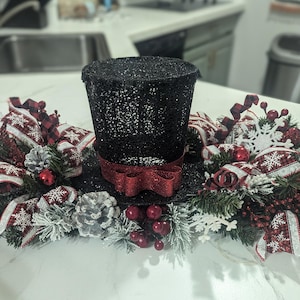 May include: A black glitter top hat with a red bow, surrounded by a festive arrangement. The arrangement includes red and white snowflake ribbons, pinecones, red berries, and faux evergreen branches, creating a holiday centerpiece.