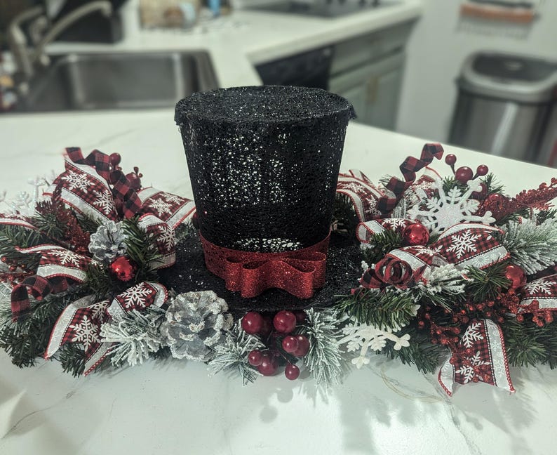 May include: A black glitter top hat with a red bow, surrounded by a festive arrangement. The arrangement includes red and white snowflake ribbons, pinecones, red berries, and faux evergreen branches, creating a holiday centerpiece.