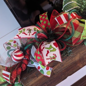 May include: A Christmas wreath with red, green, and white ribbon, featuring a green and white striped ribbon, a red and white striped ribbon, a red and white polka dot ribbon, and a ribbon with a pattern of green elves wearing red boots and holding candy canes.