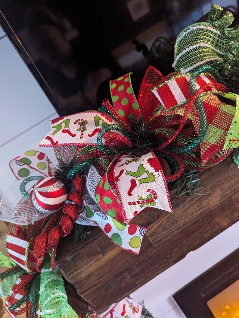 May include: A Christmas wreath with red, green, and white ribbon, featuring a green and white striped ribbon, a red and white striped ribbon, a red and white polka dot ribbon, and a ribbon with a pattern of green elves wearing red boots and holding candy canes.