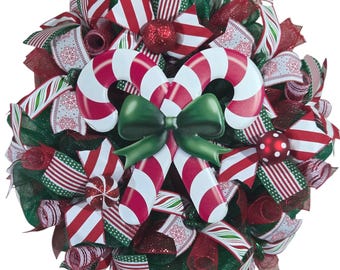 May include: A festive Christmas wreath with red, white, and green ribbons and accents. The wreath features a large candy cane design and a green bow in the center. Red glittery ball ornaments are also included.