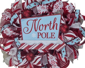 May include: A festive Christmas wreath with red, white, and light blue ribbons and ornaments. The wreath features a sign that reads "North POLE" in red lettering on a light blue background, with a snowflake graphic.