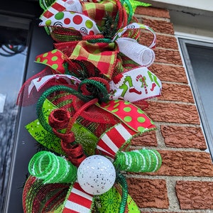 May include: A Christmas wreath with red, green, and white ribbon, a white mesh bow, and a white ornament with glitter. The wreath is decorated with a variety of Christmas-themed patterns, including snowflakes, candy canes, and polka dots.