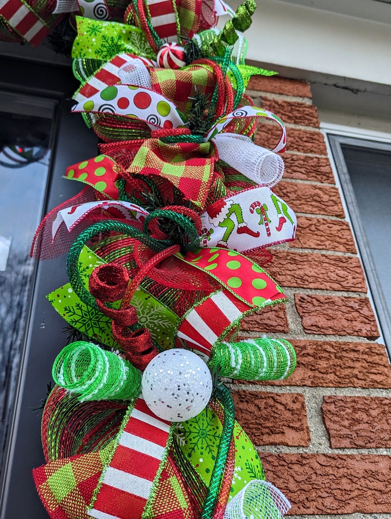 May include: A Christmas wreath with red, green, and white ribbon, a white mesh bow, and a white ornament with glitter. The wreath is decorated with a variety of Christmas-themed patterns, including snowflakes, candy canes, and polka dots.