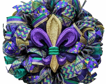 Puede incluir: Una festiva corona de Mardi Gras con un centro de flor de lis dorado. La corona está compuesta por cintas superpuestas moradas, verdes y doradas, con diseños de chevron y estampados. Un elemento decorativo.