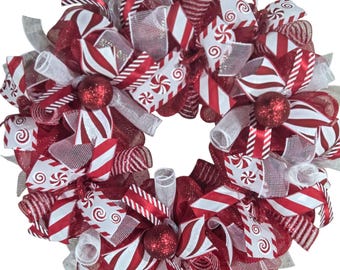 May include: A festive Christmas wreath with a candy cane theme. The wreath features red, white, and silver ribbons, bows, and mesh, adorned with red glitter ball ornaments. The design incorporates striped and swirled candy patterns.